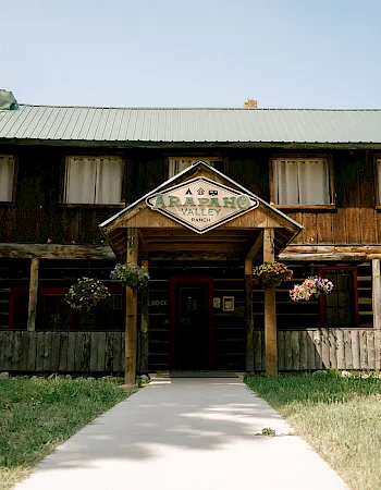 Historic wooden ranch lodge with a “Carapaho Valley Ranch” sign, flower planters, and wagon wheels in front.